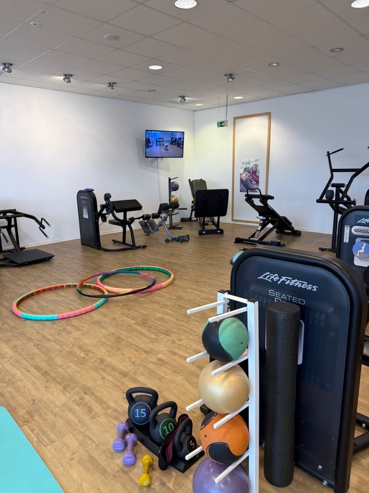 Blick über die Trainingsfläche auf ein Regal mit Medizinbällen, Kettlebells und Kurzhanteln, dahinter Ausdauergeräte und ein Bildschirm an der Wand.