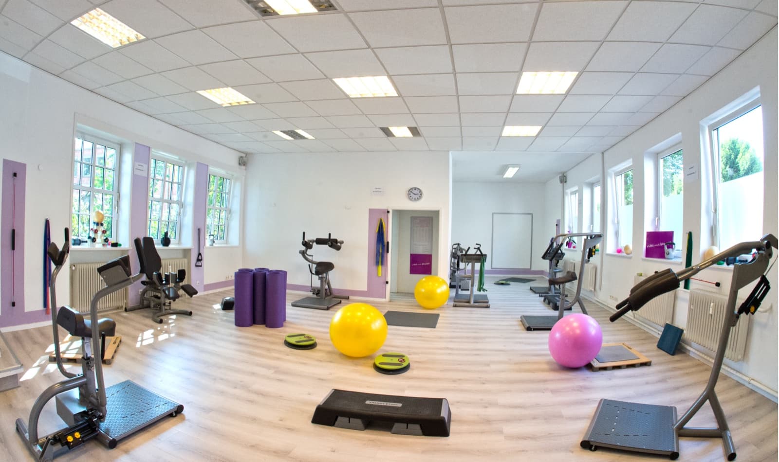 Der helle Trainingsraum mit Sprossenfenstern an beiden Seiten, gelben und rosa Gymnastikbällen, Steppbrettern und Ausdauergeräten auf hellem Holzboden.