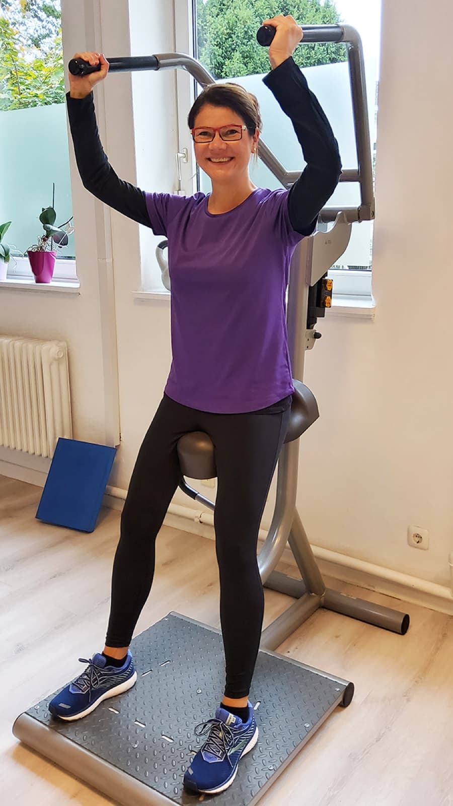 Eine Frau trainiert stehend an einem Zuggerät vor dem Fenster, unter ihr eine Vibrationsplatte, draußen Grün.