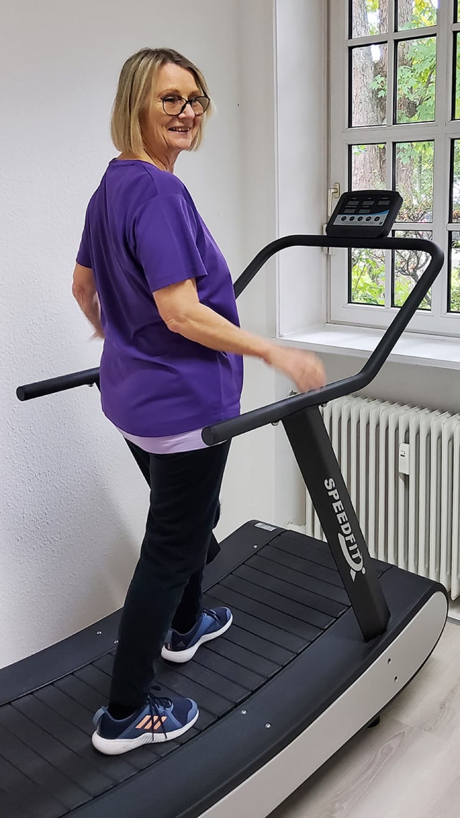 Eine Frau geht auf dem gebogenen Laufband am Sprossenfenster und dreht sich lächelnd zur Kamera.