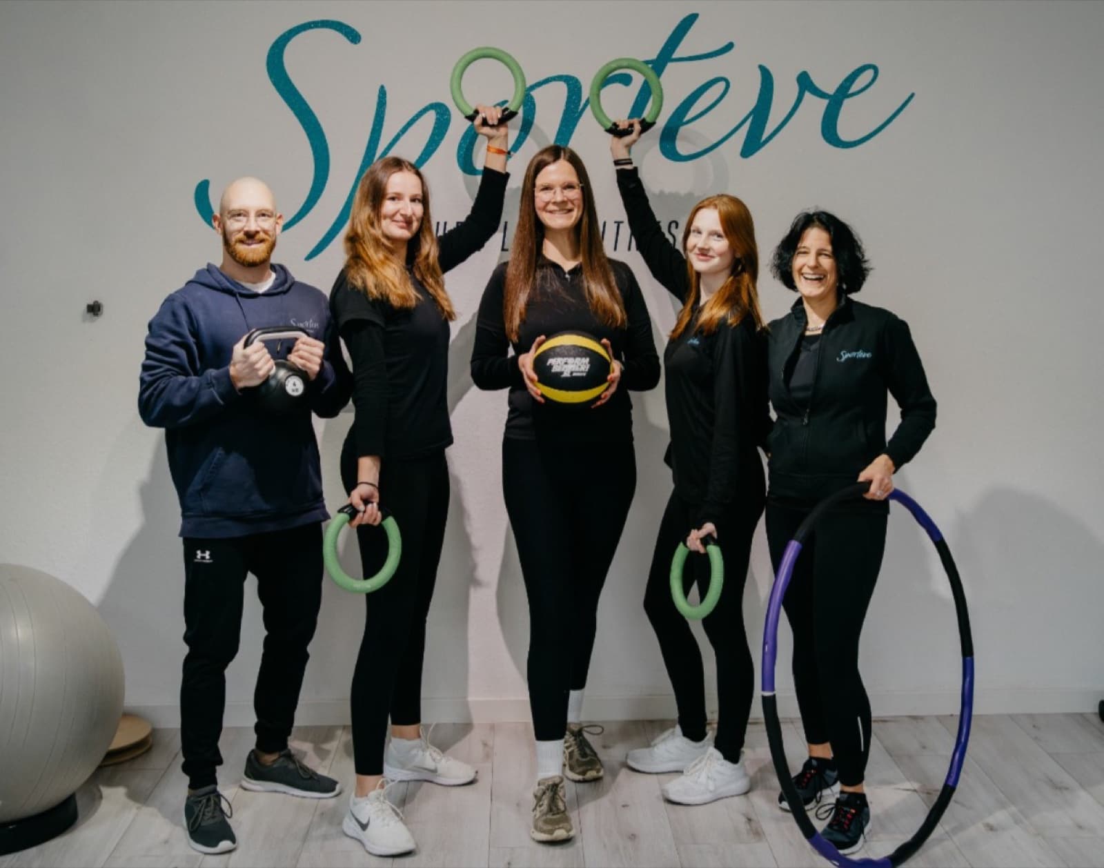 Das fünfköpfige Team vor dem Sporteve-Schriftzug an der Wand, mit Kettlebell, Gymnastikringen, Medizinball und Hula-Hoop-Reifen in den Händen.