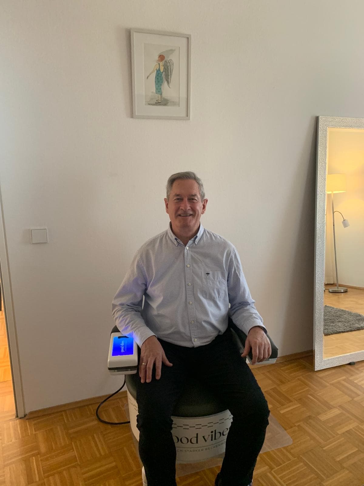 Ein Mann sitzt in Hemd und Stoffhose auf dem Beckenbodenstuhl, links daneben das beleuchtete Bedienfeld; heller Raum mit Parkett und großem Standspiegel.